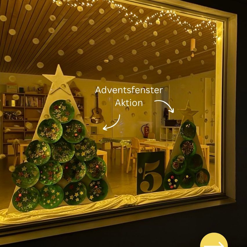 Ein Fensterdisplay mit zwei dekorierten Weihnachtsbäumen aus Papiertellern, einer mit einem Stern, und einem Schild, das Adventsfenster Aktion besagt. Hinter dem Display befindet sich ein Raum mit Tischen, Stühlen, einem Bücherregal und einer Gitarre.