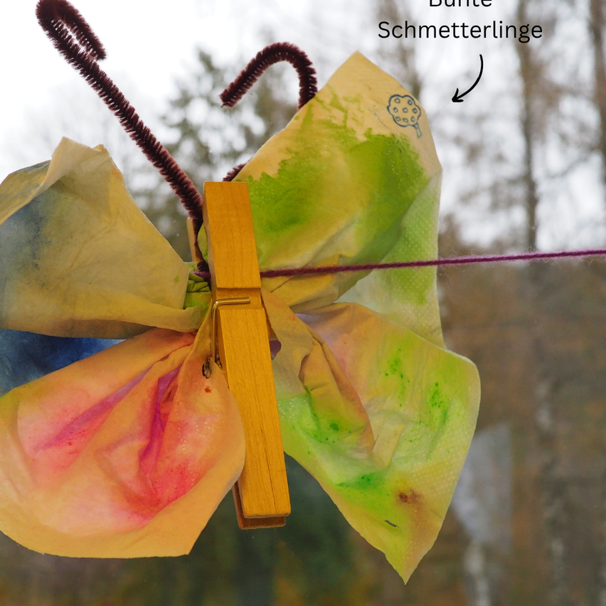 A colorful butterfly made of paper is pinned to a window with a wooden clothespin, showcasing vibrant yellow, green, and pink hues.