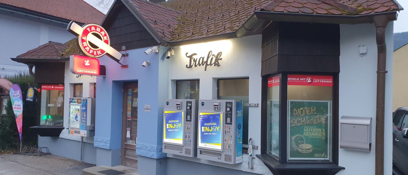 Ein Laden mit braunem Dach und blauen Wänden. Es gibt ein Schild mit der Aufschrift 'Trafik'. Zwei Maschinen sind an der Wand befestigt. Das Schild über den Maschinen steht 'Enjoy'.