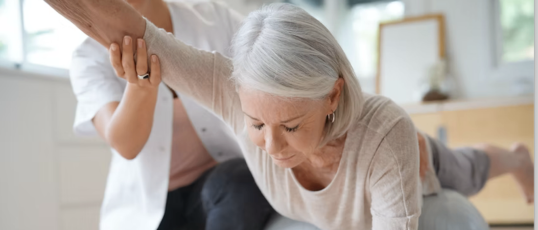 Eine ältere Frau führt unter der Anleitung einer Pflegekraft eine Übung durch. Die Frau ist über einem Trainingsball gebeugt, während die Pflegekraft sie unterstützt.