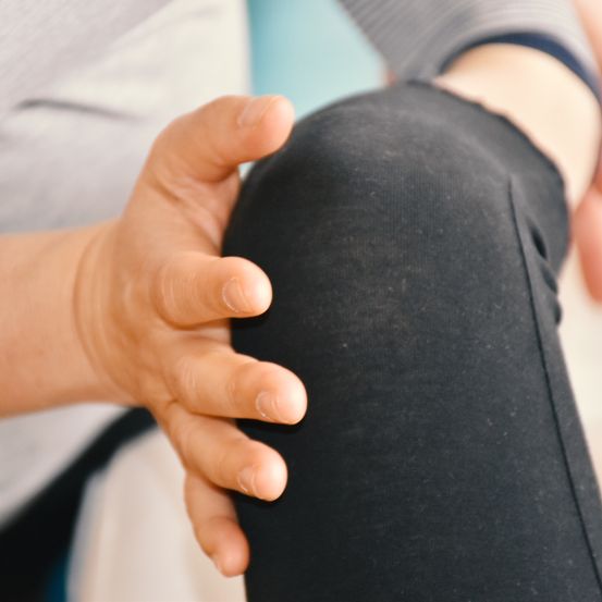 Eine Person in schwarzen Leggings hält ihr Knie mit einer Hand, wahrscheinlich in einer medizinischen Umgebung. Die Hand liegt auf der Kniescheibe, wobei die Finger sanft drücken.