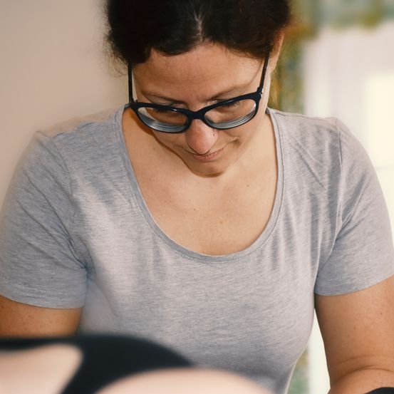 Eine Frau mit Brille beugt sich vor und scheint den Rücken einer Person zu untersuchen oder zu behandeln, möglicherweise eine Massage oder chiropraktische Behandlung.