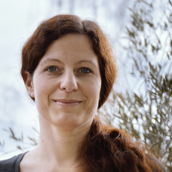 Nahaufnahme eines Porträts einer Frau mit braunen Haaren und blauen Augen, die lächelt. Sie hat einen Nasenring und steht vor einem unscharfen Hintergrund mit Olivenblättern.