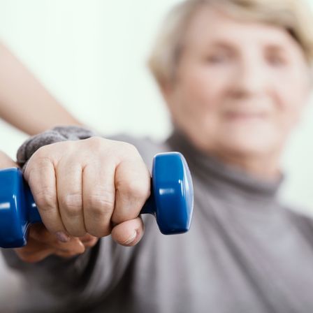 Eine ältere Frau trainiert mit einer blauen Hantel, unterstützt von jemandem, der ihre Hand hält.