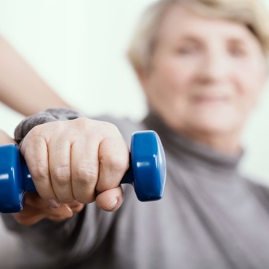 Eine ältere Frau trainiert mit einer blauen Hantel, unterstützt von jemandem, der ihre Hand hält.