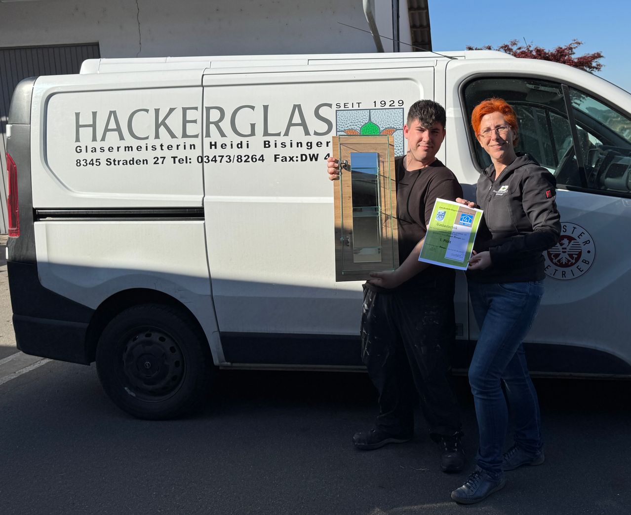 Zwei Personen stehen vor einem Van mit Hackerglas darauf. Ein Mann hält einen Holzrahmen und ein Zertifikat, während eine Frau dasselbe Zertifikat hält.