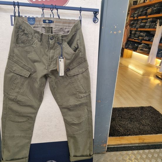 Ein Paar olivgrüne Cargo-Hosen mit mehreren Taschen hängt an einem blauen Ständer in einem Bekleidungsgeschäft, mit einem Teppich und Regalen mit gefalteter Kleidung im Hintergrund.