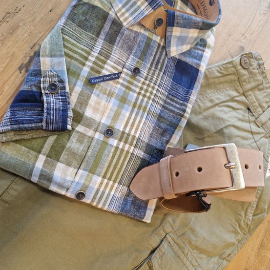 Ein kariertes Hemd und Khakihosen liegen auf einer Holzoberfläche. Das Hemd hat einen Aufnäher mit der Aufschrift 'Casual Comfort'. Ein brauner Ledergürtel liegt auf der Hose.
