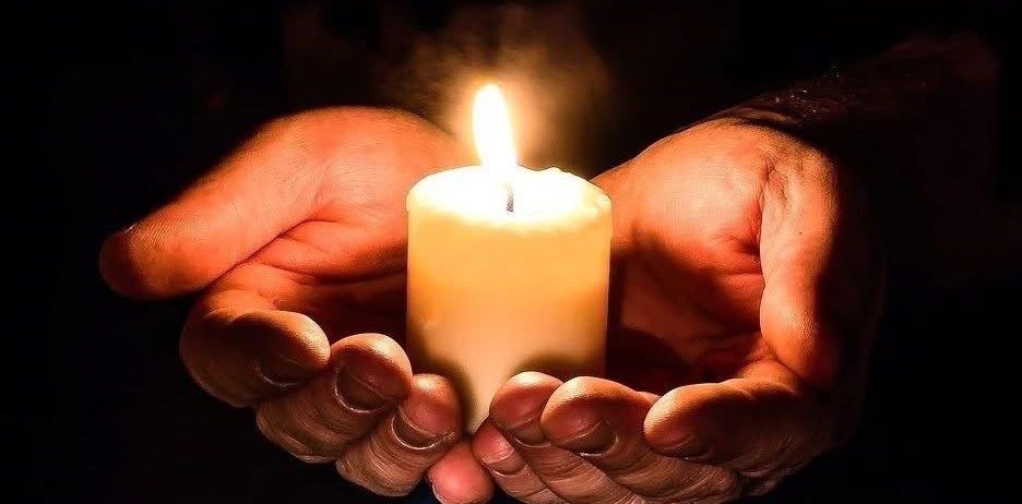 Zwei Hände halten sanft eine brennende Kerze, deren Flamme die Umgebung in einer dunklen Umgebung erhellt.