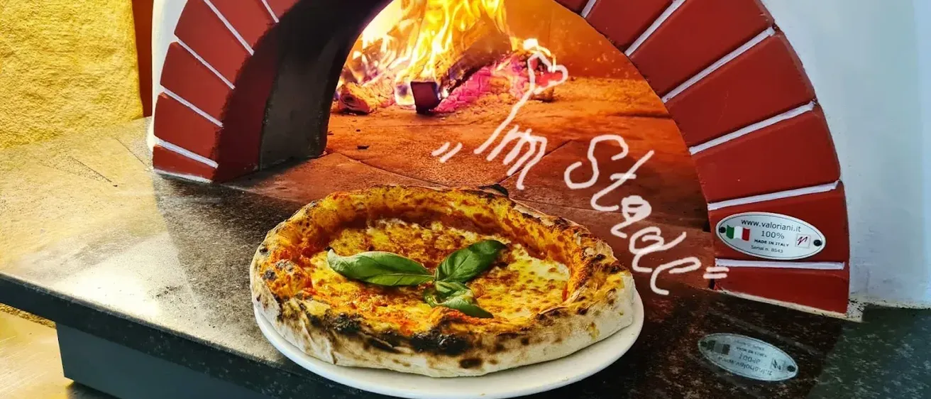 Eine frisch gebackene Pizza mit geschmolzenem Käse und Basilikumblättern auf einem weißen Teller vor einem Backofen aus Ziegelsteinen.