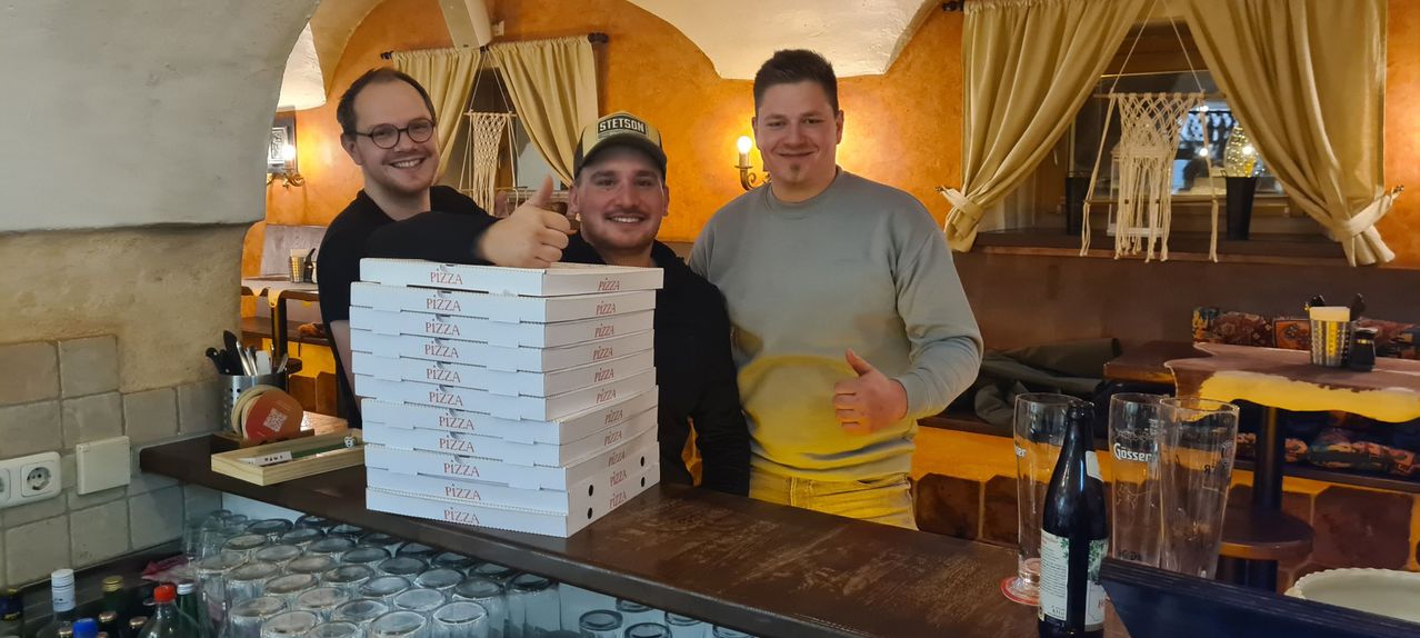 Drei Männer stehen hinter einer Theke. Sie lächeln und geben Daumen hoch. Ein Stapel Pizzakartons steht vor ihnen. Ein Glas steht auf der Theke.