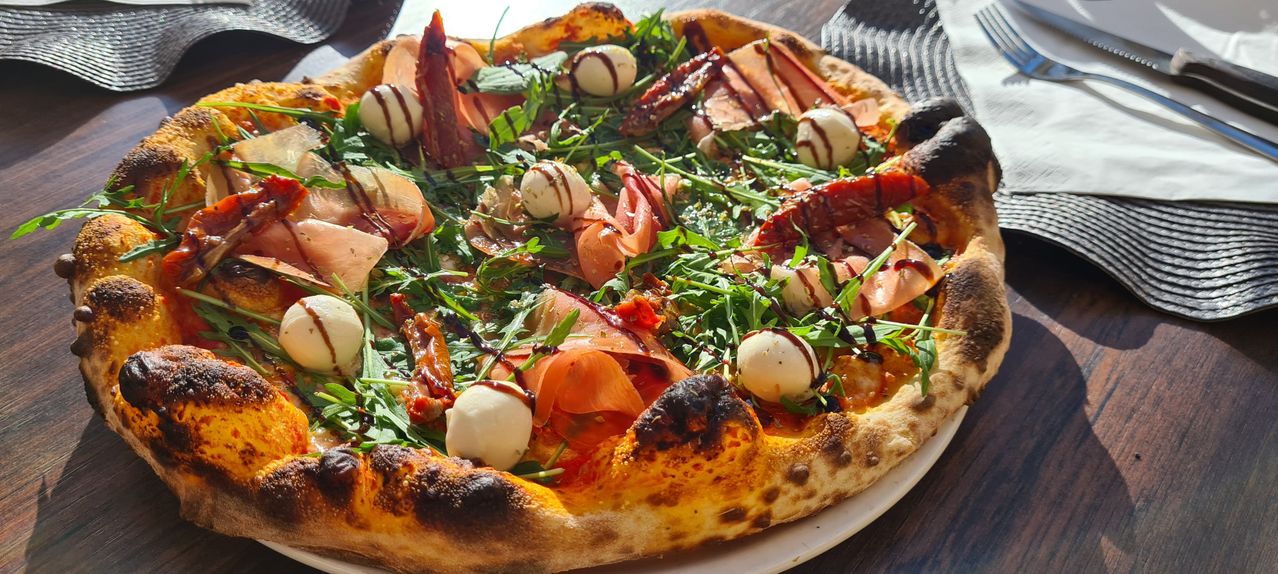 Eine Pizza mit Rucola, Kirschtomaten, Mozzarellabällchen und Prosciutto-Scheiben, leicht mit Sauce überzogen.