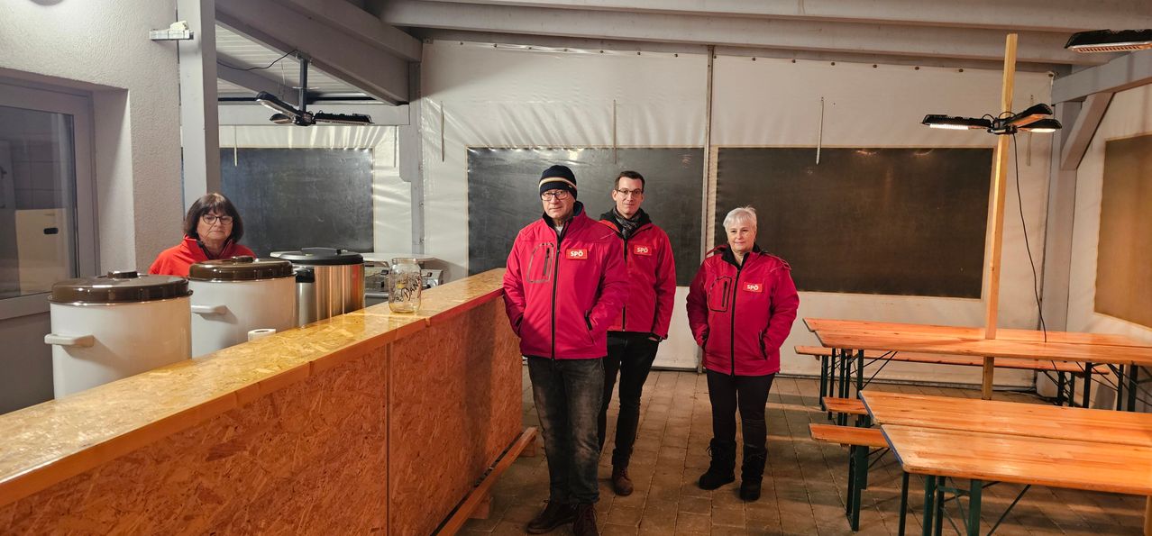 Drei Personen in roten Jacken stehen in einem Raum mit weißen Wänden und einem gefliesten Boden. Ein Tresen und ein Tisch mit Bänken sind vorhanden. Ein Glaskrug steht auf dem Tresen.