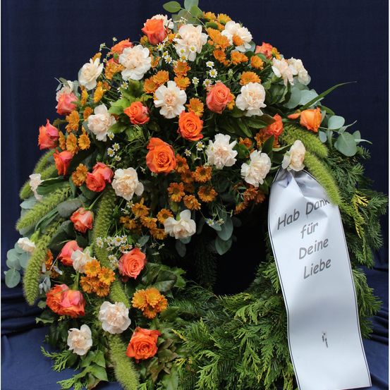 Ein Kranz aus orangefarbenen und weißen Blumen mit einem Band, das 'Danke für deine Liebe' auf Deutsch sagt. Die Blumen sind in einem Kreis mit blauem Hintergrund angeordnet.