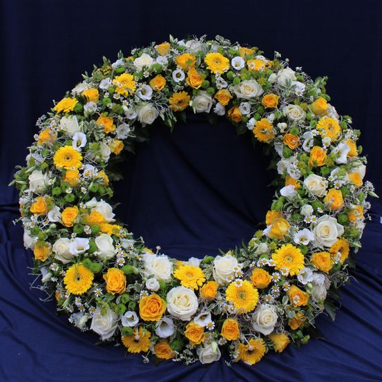 Ein kreisförmiger Blumenkranz aus weißen, gelben und grünen Blumen wird auf einem blauen Hintergrund präsentiert. Die Blumen sind in einer kreisförmigen Form angeordnet.