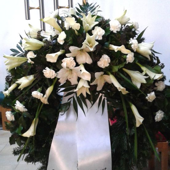 Ein weißer Blumenkranz mit verschiedenen weißen Blüten, einschließlich Lilien und Nelken, ist ausgestellt. Der Kranz ist mit grünen Blättern verziert und hat ein weißes Band darum geschlungen. Er steht auf einer weißen Oberfläche, möglicherweise einem Tisch oder Altar.