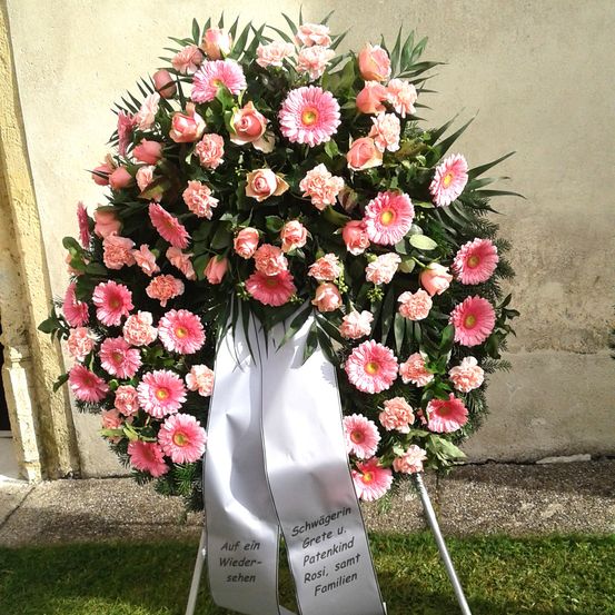 Ein Kranz aus rosa Blumen mit einer weißen Schleife, auf Gras platziert. Eine Nachricht in Deutsch ist auf der Schleife, mit einem Hintergrund eines Gebäudes.