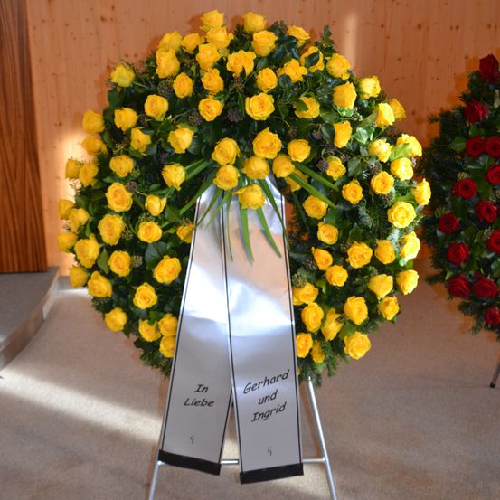 Ein gelber Kranz mit silbernen Bändern, auf denen 'In Liebe, Gerhard und Ingrid' steht, wird in einem Raum ausgestellt.