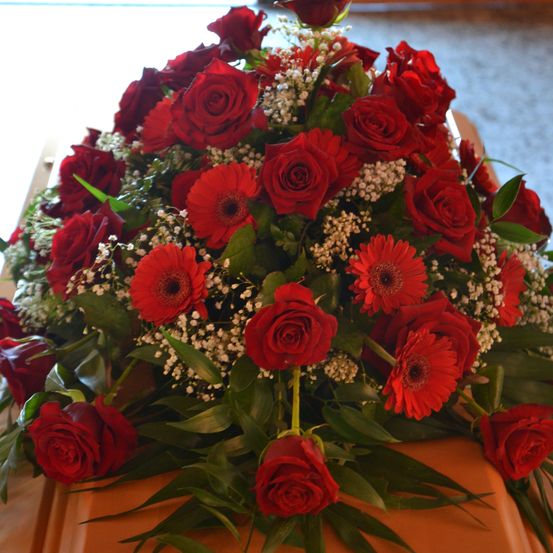 Ein Sarg bei einer Beerdigung, geschmückt mit einem Strauß roter Rosen, Babysatem und roten Gerbera-Daisies auf einer Holzoberfläche.