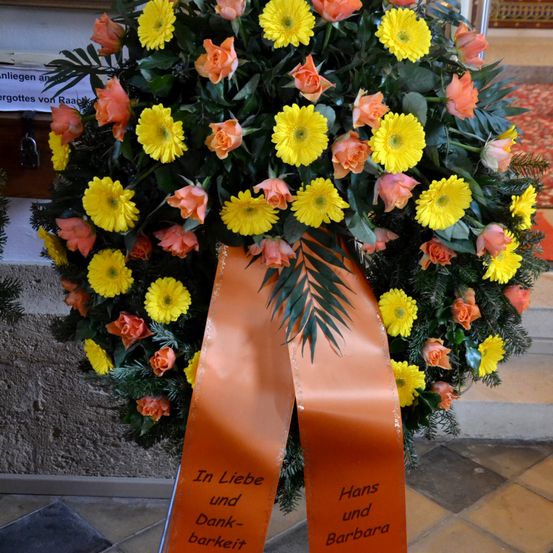 Ein Kranz mit gelben und orangefarbenen Blumen, geschmückt mit grünen Blättern, zeigt ein orangefarbenes Band mit der Aufschrift 'In Liebe und Dankbarkeit.' Es handelt sich wahrscheinlich um einen Gedenkkranz.