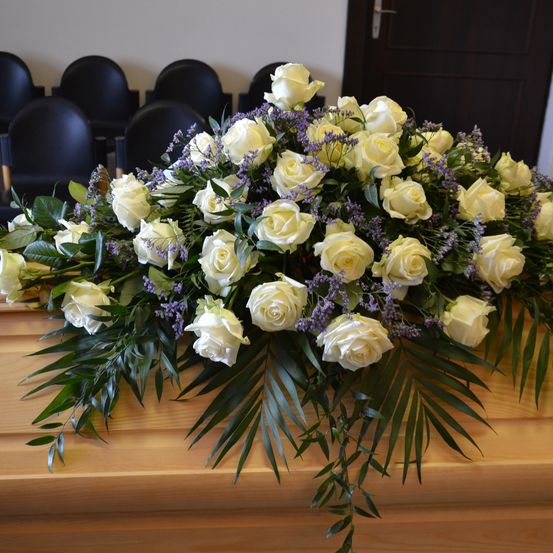 Eine große Blumenarrangement aus weißen Rosen und lila Blumen auf einem hölzernen Sarg, in einem Raum mit schwarzen Stühlen platziert.