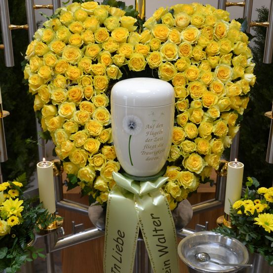 Ein herzförmiger Strauß gelber Rosen umgibt eine weiße Urne mit einem Löwenzahn darauf. Ein Band mit der Aufschrift 'In Liebe' ist zu sehen. Kerzen und Grünpflanzen umgeben die Urne.