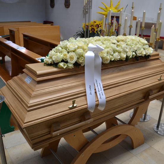 Ein hölzerner Sarg mit weißen Rosen und einer Schleife mit der Aufschrift 'In Liebe' steht in einer Kirche mit gefliestem Boden und Kerzenständern.