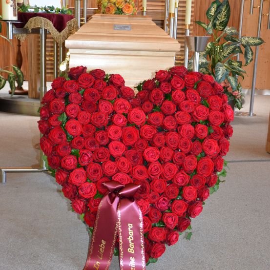 Ein herzförmiges Arrangement aus roten Rosen steht vor einem hölzernen Sarg. Der Sarg steht auf einer Plattform mit einem roten Band, auf dem 'In Liebe' steht. Dahinter befindet sich ein Kerzenständer und eine Pflanze.