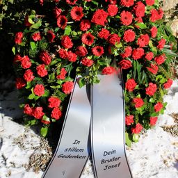 Ein Kranz aus roten Blumen und grünen Blättern auf schneebedecktem Boden. Zwei graue Bänder mit schwarzem Text lauten 'In stillem Gedenken' und 'Dein Bruder Josef'.