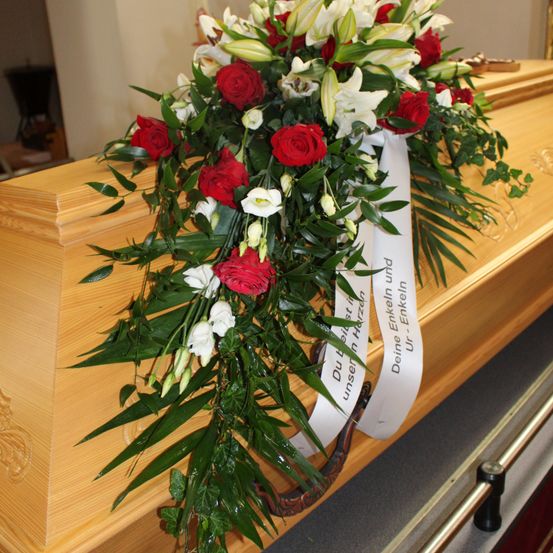 Ein hölzerner Sarg, geschmückt mit einer Blumenanordnung aus roten und weißen Blumen, grünen Blättern und einer weißen Schleife mit deutschem Text.