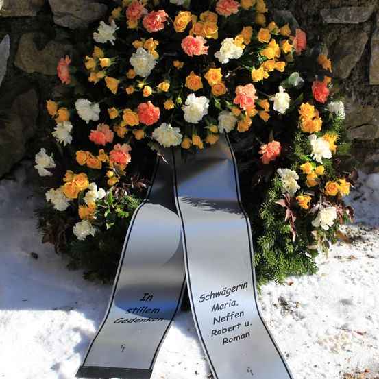 Ein Kranz mit orangen, weißen und gelben Blumen, umgeben von Grün, liegt auf einer schneebedeckten Oberfläche. Ein grauer Schriftband mit dem deutschen Text 'In stillem Gedenken' ist befestigt.