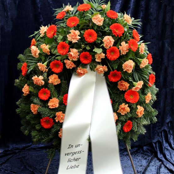 Ein Kranz mit orangefarbenen Blumen und grünen Blättern hat ein weißes Band mit schwarzem Text, der 'In Unvergesslicher Liebe' lautet. Er ist auf einem blauen Hintergrund ausgestellt.