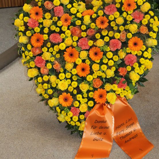 Ein herzförmiger Blumenstrauß mit gelben, orangen und roten Blumen. Ein orangefarbenes Band mit einer auf Deutsch geschriebenen Botschaft ist am Strauß befestigt.