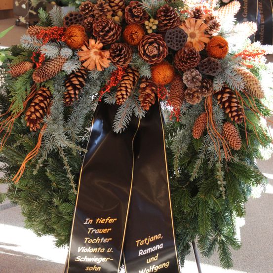 Ein Kranz aus Tannenzapfen und grünen Zweigen mit einem schwarzen Band, auf dem steht 'In tiefer Trauer Tochter Violanta u. Schwieger-sohn'.