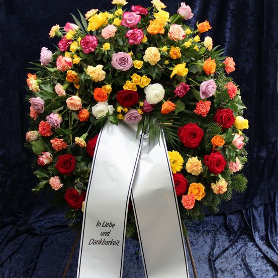 Ein Kranz mit verschiedenen farbigen Rosen und Nelken, umwickelt mit einem weißen Band mit schwarzen Rändern. Er steht auf einem blauen Hintergrund. Das Band trägt den Schriftzug 'In Liebe und Dankbarkeit'.