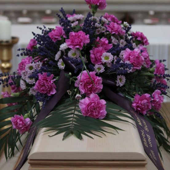 Ein Sarg mit einem lila Band und Blumenarrangement aus rosa und blauen Blumen, einschließlich Nelken und Lavendel, auf einer weißen Oberfläche.