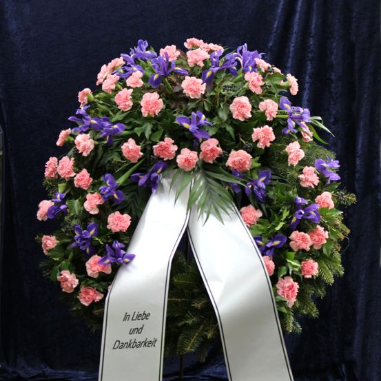 Ein Kranz aus rosa Nelken und lila Schwertlilien mit einer weißen Schleife, auf der 'In Liebe und Dankbarkeit' steht, vor einem dunkelblauen Hintergrund.
