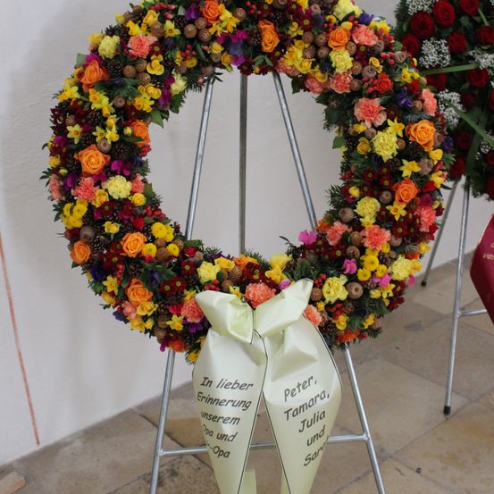 Ein farbenfroher Kranz aus verschiedenen Blumen, einschließlich Rosen, Nelken und Gänseblümchen, wird auf einem Ständer an einer weißen Wand ausgestellt. Er enthält ein weißes Band mit schwarzem Text.