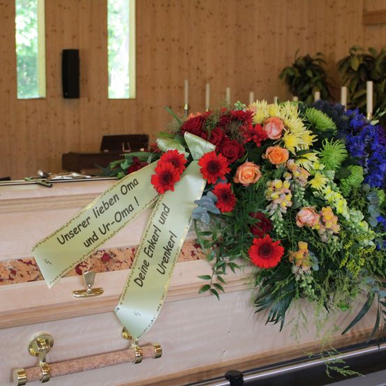 Ein Sarg mit einem Strauß bunter Blumen und einer Schleife mit dem Text 'Unserer lieben Oma und Ur-Oma!' steht auf einer Holzoberfläche. Dahinter befindet sich eine Holzwand mit Fenstern und einem Lautsprecher.