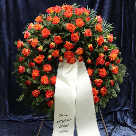 Ein Kranz aus roten und orangefarbenen Rosen mit grünen Blättern und einer weißen Schleife mit dem Text 'In unvergesslischer Liebe' wird vor einem blauen Hintergrund gezeigt.