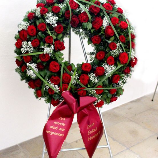 Ein Kranz aus roten Rosen mit einem roten Band steht auf einem Metallständer. Das Band hat goldene Schrift, die 'Ich werde dich nie vergessen' liest. Der Hintergrund ist eine helle Wand.