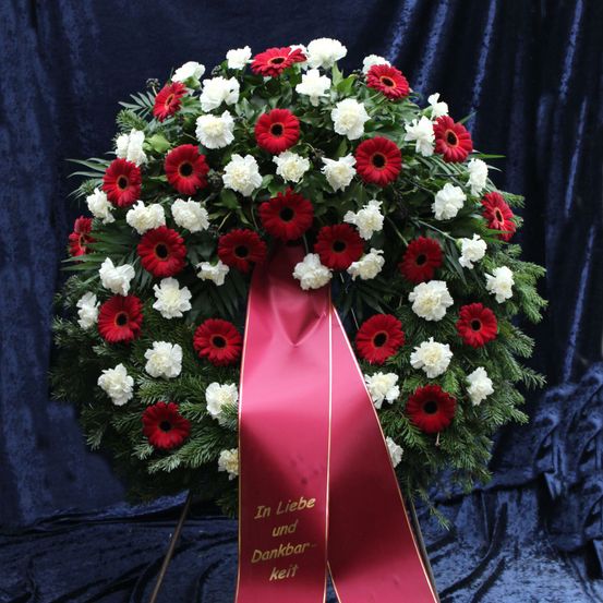 Ein Kranz mit roten und weißen Blumen auf blauem Hintergrund, geschmückt mit einem roten Band, das in goldenen Buchstaben 'In Liebe und Dankbarkeit' trägt.