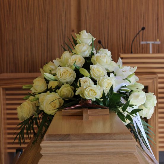 Ein mit weißen Rosen und Lilien geschmückter Sarg steht in einer Kapelle, ein hölzerner Hintergrund dahinter, ein Kreuz und Mikrofone sichtbar.