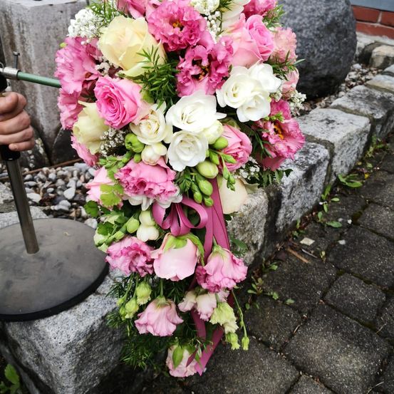 Ein großer Strauß aus rosa und weißen Blumen mit einem rosa Band wird von einer Hand gehalten und auf einer Steinoberfläche platziert.