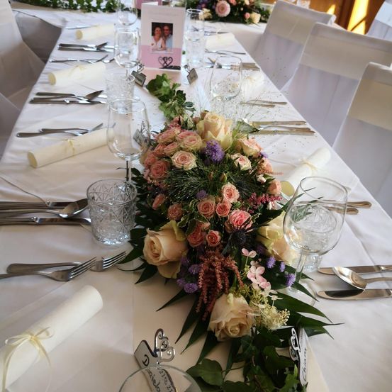 Ein langer Esstisch, der für einen besonderen Anlass gedeckt ist. Es gibt einen Blumen-Zentralteil, Weingläser und Besteck. Weiße Servietten, ein rosa Fotorahmen und Stühle sind ebenfalls vorhanden.