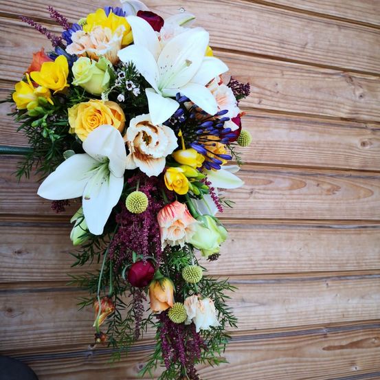 Ein farbenfroher Strauß aus verschiedenen Blumen, einschließlich weißer Lilien, gelber, oranger und rosa Rosen und anderen bunten Blüten, hängt an einer Holzwand.