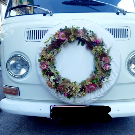 Ein weißer VW-Bus zeigt einen Blumenkranz auf dem Stoßfänger. Der Kranz besteht aus verschiedenen Blumen und Grün.