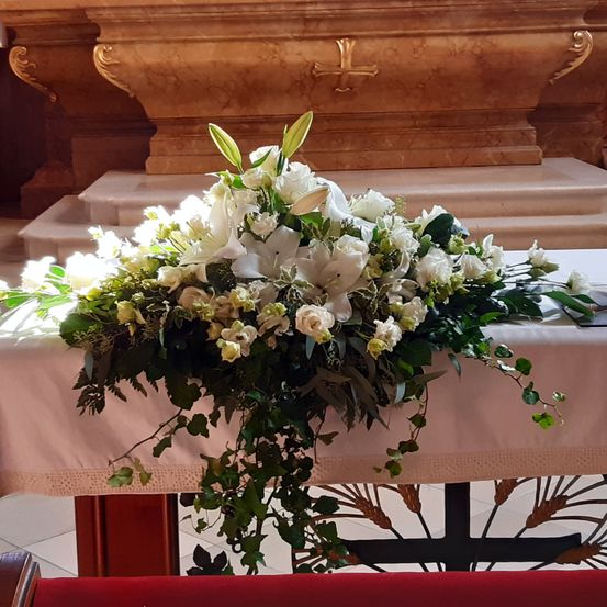 Eine weiße Blumenarrangement mit Lilien und Grünpflanzen steht auf einem weißen Tisch in einer Kirche. Dahinter befindet sich ein Marmoraltar mit einem Kreuz.