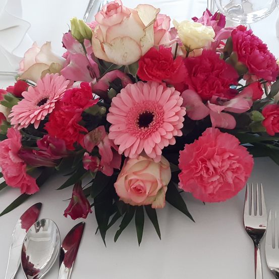 Eine pinke Blumenarrangement mit Rosen, Gänseblümchen und Nelken auf einer weißen Tischdecke. Silberne Bestecke und ein Glas sind in der Nähe.