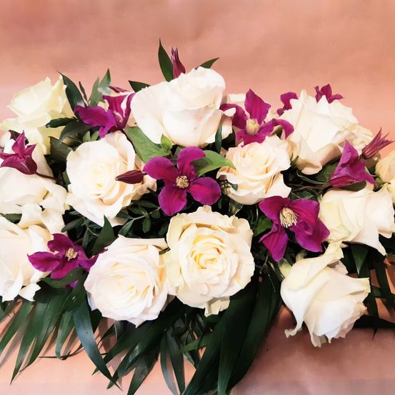 Ein Blumenarrangement mit weißen Rosen und lila Blumen mit grünen Blättern auf einem rosa Hintergrund.